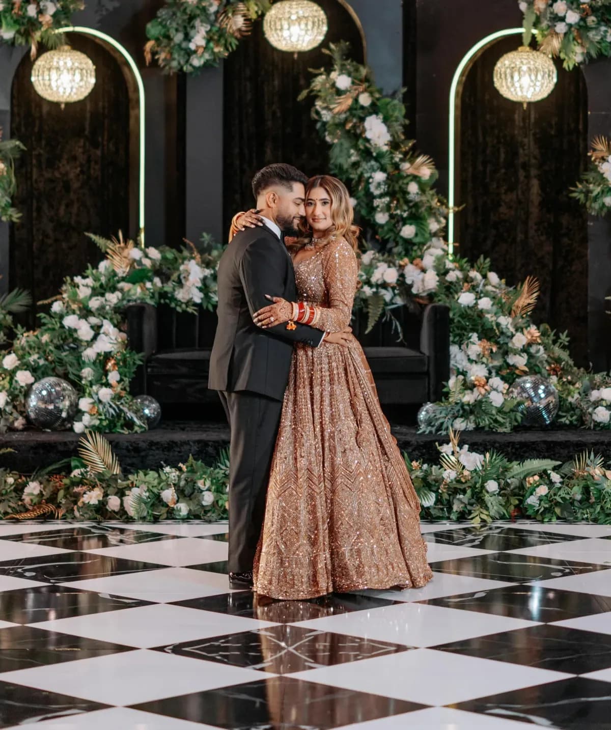 Couple dancing on elegant black and white checkered dance floor wrap with crystal chandeliers and lush floral backdrop at luxury wedding reception